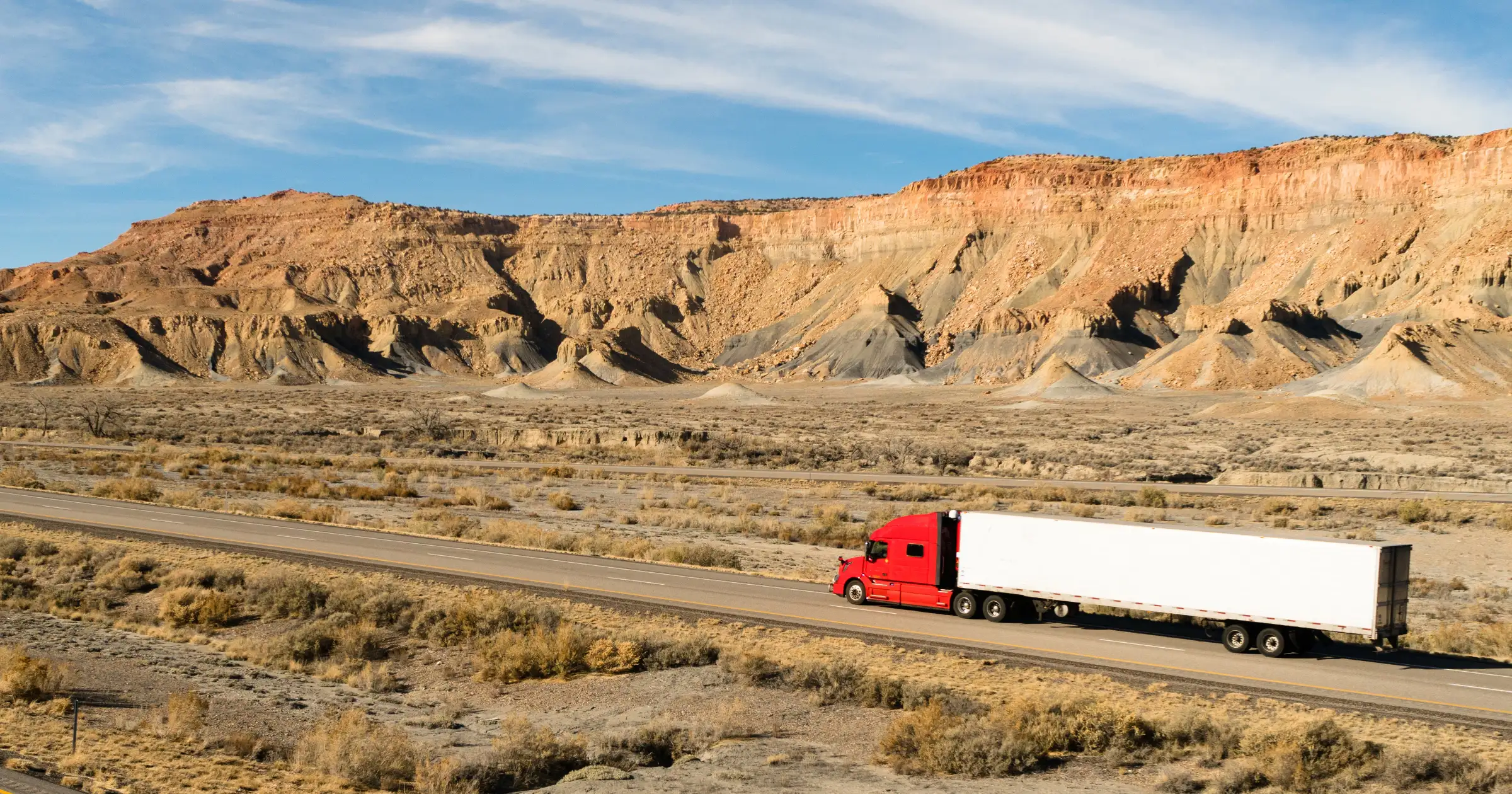 Red semi-truck with a white trailer driving along a desert highway with rocky cliffs and mesas in the background.
