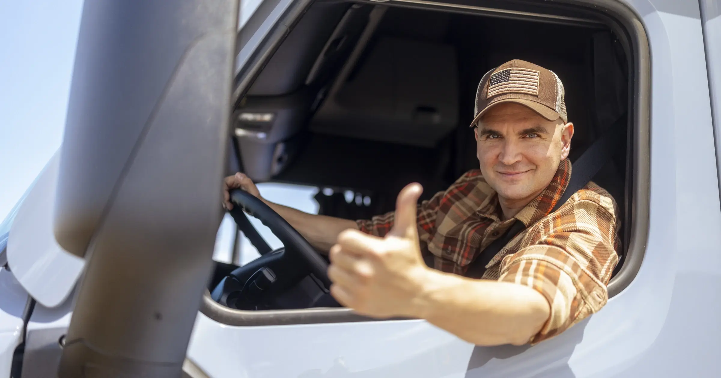 A truck driver wearing a plaid shirt and a cap sits in the cab of his semi-truck and gives a thumbs-up toward the camera with a smile.