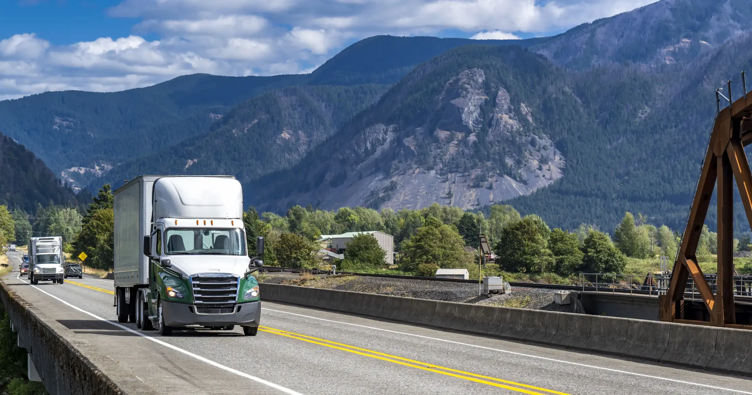 Semi-trucks driving along a mountain highway with a scenic backdrop of forested hills and a cloudy blue sky.