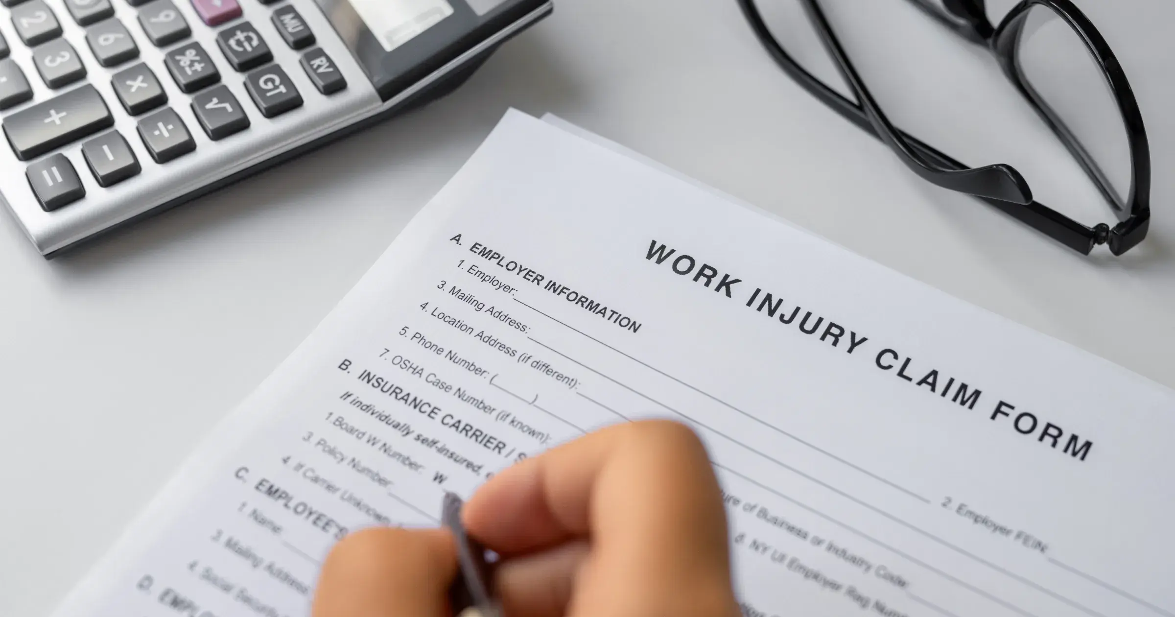 Person filling out a work injury claim form at a desk with a calculator and eyeglasses, representing workers’ compensation insurance paperwork.