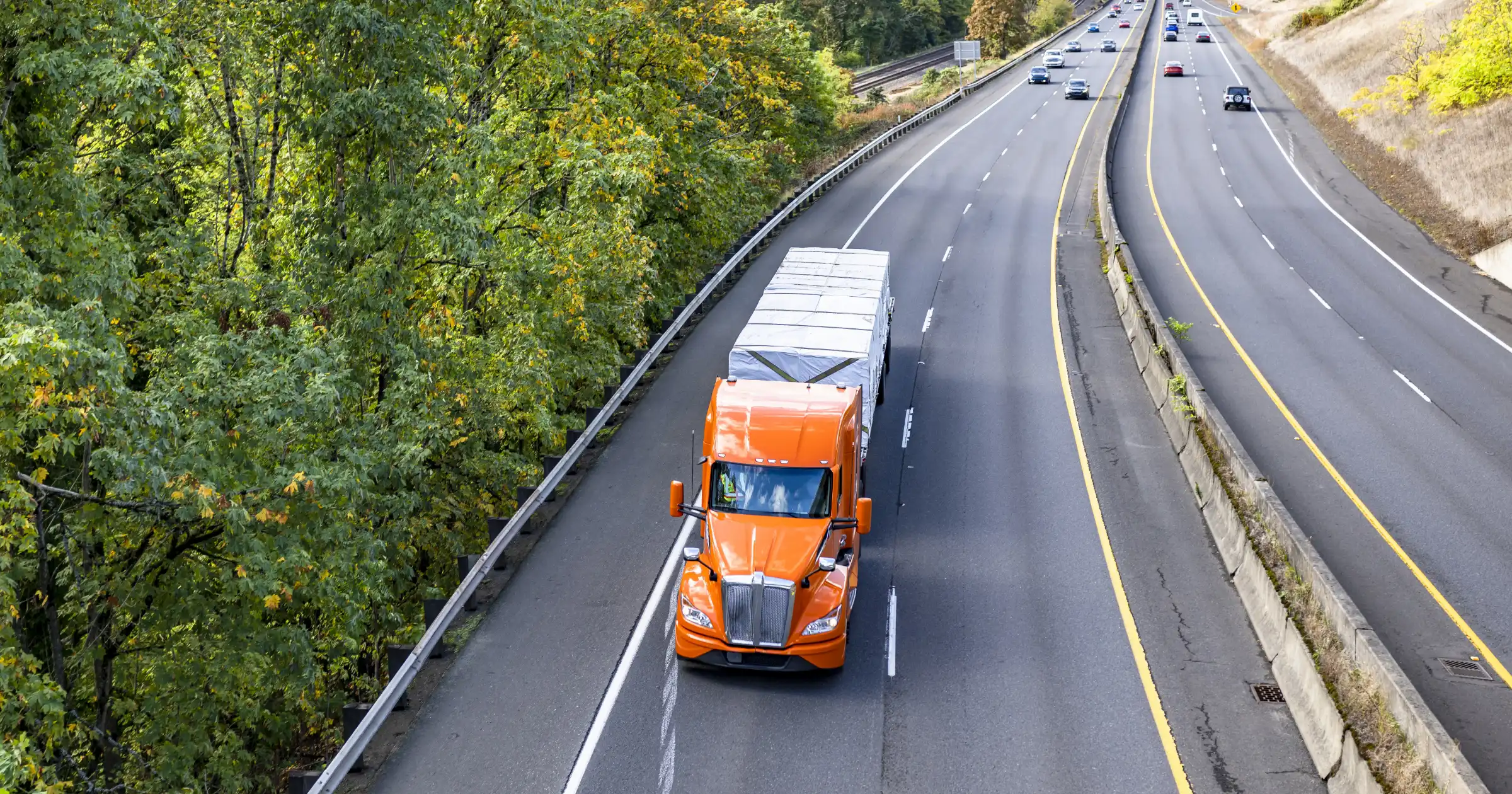 An orange semi-truck hauling a covered load drives down a highway surrounded by trees on one side and a concrete median separating traffic lanes.